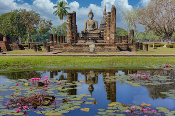 Fototapeta na wymiar View of ruins of Wat Mahathat temple in Sukhothai historical park with sitting Buddha and reflection on pond with pink water lilies. Travel concept.