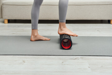 Cropped view of young barefoot woman in sportswear massaging feet with roller massager and standing on fitness mat at home, promoting lymph flow and wellness at home concept, tension relief