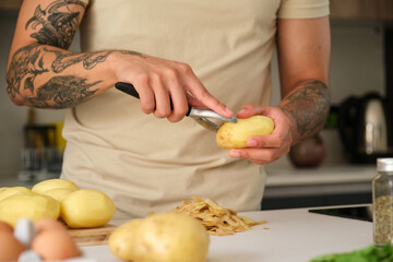 Young unrecognizable tattooed latin man peeling potatoes at kitchen to prepare a recipe.