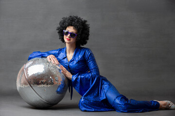 Spectacular Woman in a sparkly flared suit and afro wig poses with a disco ball. Disco style from...