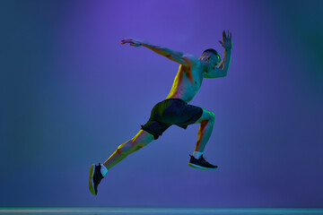 Fototapeta na wymiar Shirtless man, with fit, relief, muscular body, professional athlete in motion, running against blue studio background in neon light