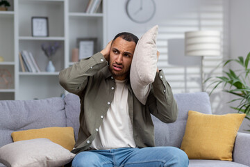 A young African American man is sitting on the sofa at home and covers his ears with pillows. Angry...