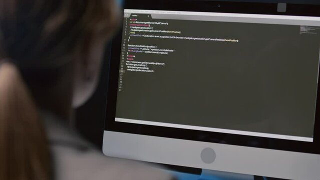 Over-the-shoulder close up of female programmer sitting at desk writing code in office at night