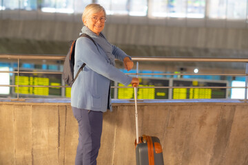 Smiling senior traveler woman with trolley and backpack walking through airport area to reach departure gate. Old senior woman leaving for vacation