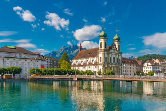 Picturesque view of the Lucerne Jesuit Church St. Francis Xavier at the river Reuss with the bridge Rathaussteg in front and the mountains in the back on a sunny day with a blue sky.