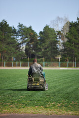 A man on a lawn mower mows the grass. The stadium has been cleared of vegetation. A field for sports games.