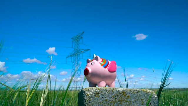 Una persona echando monedas en una hucha de cerdito unicornio en un campo de trigo con torres el&eacute;ctricas a su espalda como concepto del precio de la electricidad, ahorro energ&eacute;tico, medioambiente, etc