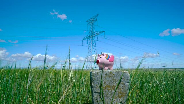 Una persona echando monedas en una hucha de cerdito unicornio en un campo de trigo con torres el&eacute;ctricas a su espalda como concepto del precio de la electricidad, ahorro energ&eacute;tico, medioambiente, etc