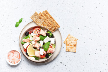 Fresh salad made with tomatoes, olives, cucumbers, onion, cheese and roasted salmon in the ceramic bowl  and full grain crackers on white marble background top view, copy space for your design.