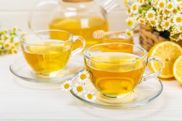 Chamomile herbal tea in a glass cup on a white wooden table with honey, lemon and chamomile bouquet. Close-up. Copy space. Useful herbal drinks, immunity tea. Natural healer concept.Place for text.