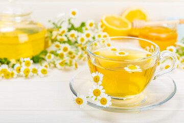 Chamomile herbal tea in a glass cup on a white wooden table with honey, lemon and chamomile bouquet. Close-up. Copy space. Useful herbal drinks, immunity tea. Natural healer concept.Place for text.