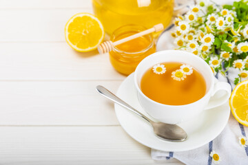 Herbal chamomile tea in a white cup on a white wooden table with honey, lemon and chamomile bouquet. Close-up. Copy space. Useful herbal drinks, immunity tea. Natural healer concept.