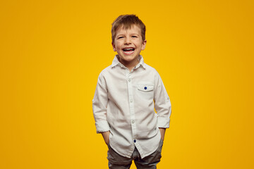 Cute little boy in trendy white shirt smiling broadly while keeping hands in pockets against yellow background