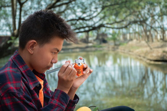 Young asian boy in plaid shirt sitting by the pond in the backyard and spending his free time by drawing, coloring and fixing his fruit and animal models sculpted from plaster with watercolor happily.