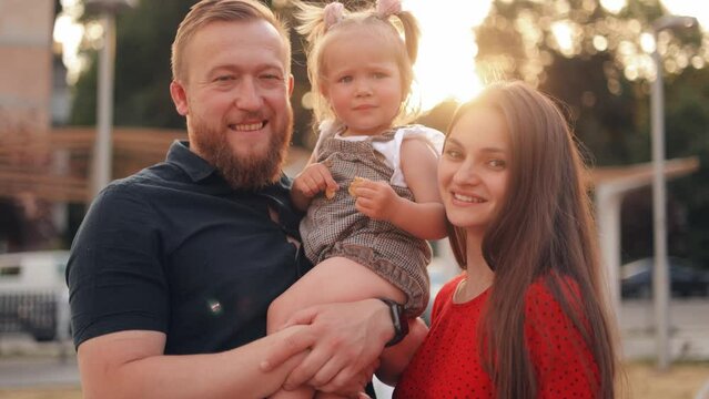 Happy attractive young mom and dad holding sweet little daughter girl in arms, looking at camera, away with toothy smiles, laughing, hugging enjoying leisure, close family relationships