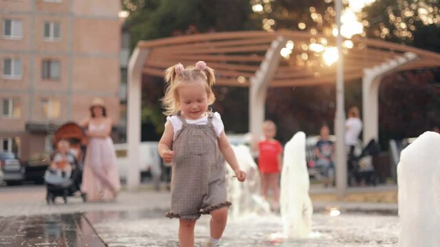 Happy little girl is playing with the stream of water, running on the water. A child is playing near a fountain. Fountain in the town square outdoors. Summer vacation and rest
