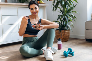 Athletic woman eating a healthy bowl of muesli with fruit sitting on floor in the kitchen at home