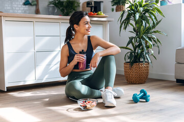 Athletic woman drinking healthy protein shake while sitting on floor in the kitchen at home