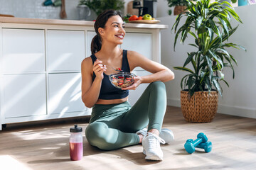 Athletic woman eating a healthy bowl of muesli with fruit sitting on floor in the kitchen at home - obrazy, fototapety, plakaty
