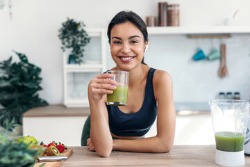 Athletic smiling woman drinking smothie while listening music with earphones in the kitchen at home.