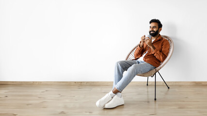 Cheerful indian man drinking coffee while chilling in armchair indoor