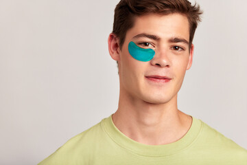 A millennial guy with brown hair practicing skincare with under eye hydrogel patches in a studio. Young caucasian man smiling, absorbing the ingredients of the cosmetic under eye mask. Men's skincare.