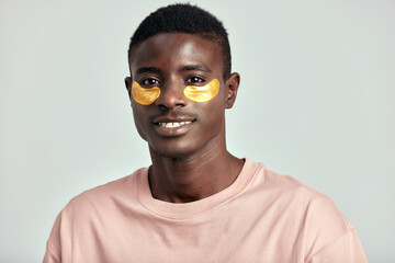 African millennial guy practicing skincare and grooming with under eye hydrogel patches in a studio. Young black man smiling, absorbing the ingredients of the cosmetic under eye mask. Men's skincare.