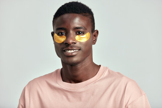 African millennial guy practicing skincare and grooming with under eye hydrogel patches in a studio. Young black man smiling, absorbing the ingredients of the cosmetic under eye mask. Men's skincare. - Powered by Adobe