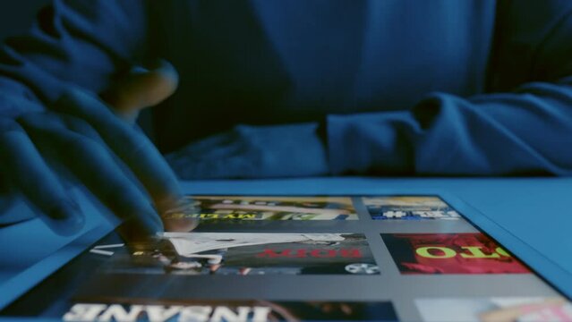 Timelapse close up of unrecognizable woman sitting at table with tablet on it choosing film to watch on streaming service at night time