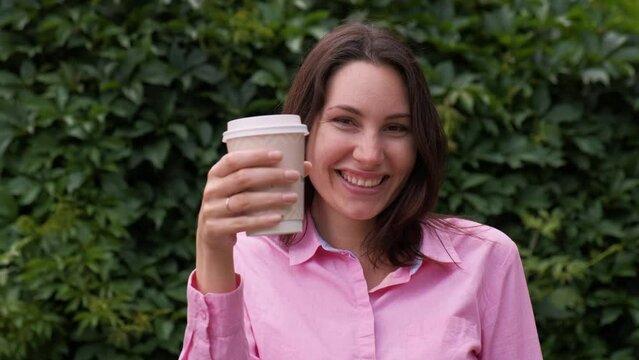 An attractive woman looks into the camera, raises a paper cup with cappuccino and smiles against the background of a grass fence. Coffee advertising