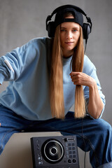 A young pretty long-haired DJ girl in a blue sweater, jeans, a black baseball cap and black headphones sits with a DJ mixing console. Studio shot, gray background.