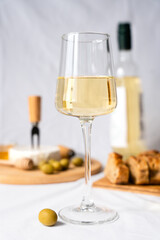 A wineglass of white wine and an olive on the table. In the background in blur a bottle of wine, camembert cheese, baguette and olives. Selective focus.
