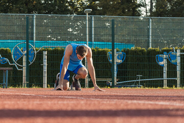 Rear view of a male Caucasian sprinter assuming a starting position in the starting block during running training at an athletic track, ground level close up shot.