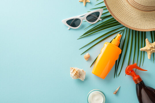 Safe beach tan concept. High angle view photo of straw hat, sunglasses, marine shells and sunscreen products on palm leaf on light blue isolated background with copy-space