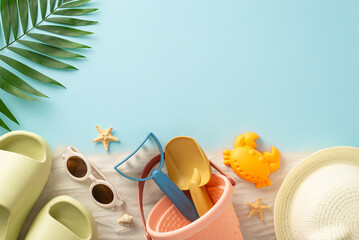 Beach relaxation on the seaside with children. Top view photo of sand with sand toys for kids, panama, slippers, sunglasses, palm leaf and starfish on isolated blue background with copyspace