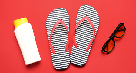 Sunglasses, flip-flops and bottle of sunscreen cream on red background