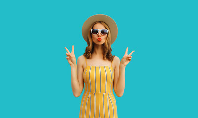 Portrait of beautiful young woman posing blowing her lips sending sweet air kiss wearing summer straw hat and dress on blue background