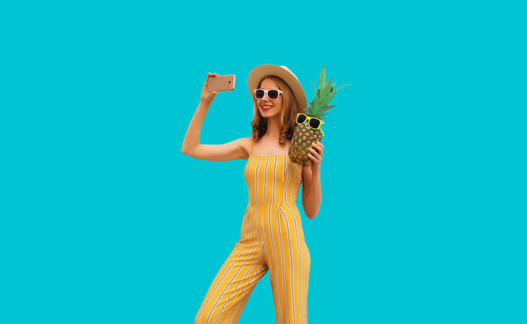 Summer look, stylish happy smiling woman holding pineapple taking selfie with smartphone wearing straw hat, jumpsuit and sunglasses on blue background