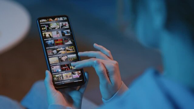 Panning left close up of unrecognizable woman sitting on sofa holding smartphone, looking at screen with streaming service and choosing film