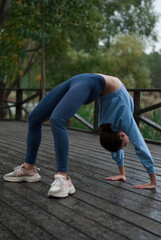 View of a girl with athletic body in blue gym suit is doing exercises in rainy weather in the park.