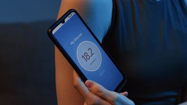Close up of unrecognizable woman with electronic patch on shoulder checking glucose level with special app on smartphone indoor at night time