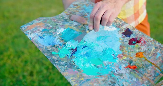 The artist paints a picture in the park. Green landscape, grass trees and flowers. The easel on which the painting stands. Blue, green colors in the palette.