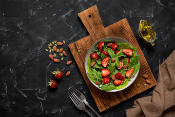 Fototapeta na wymiar Summer vegan salad with strawberries, arugula, spinach, nuts and pumpkin seeds in a bowl on a black stone background. Top view, flat lay, copy space