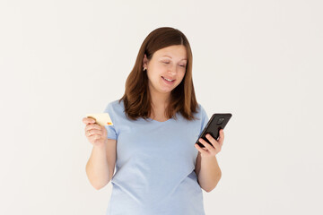 Online shopping. Portrait of smiling pregnant woman paying with plastic credit card on smartphone app, standing with mobile phone and bank card against white background