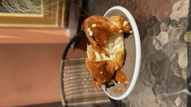 Close-up of a delicious cruffin with salted caramel torn into halves, caramel dripping down, on a saucer at a glass table of a cafe terrace. Lifestyle vertical video.
