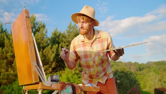 The artist paints a picture in the park. Green landscape, grass trees and flowers. The easel on which the painting stands. Blue, green colors in the palette.