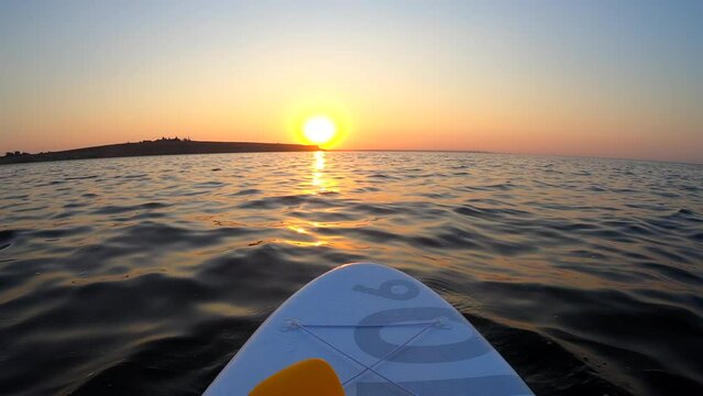 White SUP board with yellow oar on surface of sea at sunset dawn. Person swims on stand-up paddleboard in ocean near shore at sunrise sunset. POV. Wide below angle. Active lifestyle sport recreation