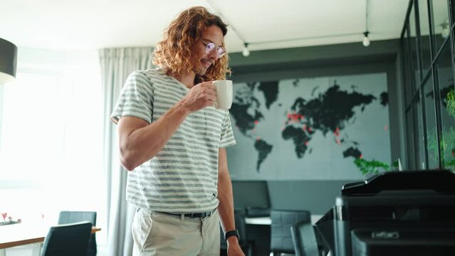 Young curly long haired man in eyeglasses drinking coffee and prints documents on printer in office