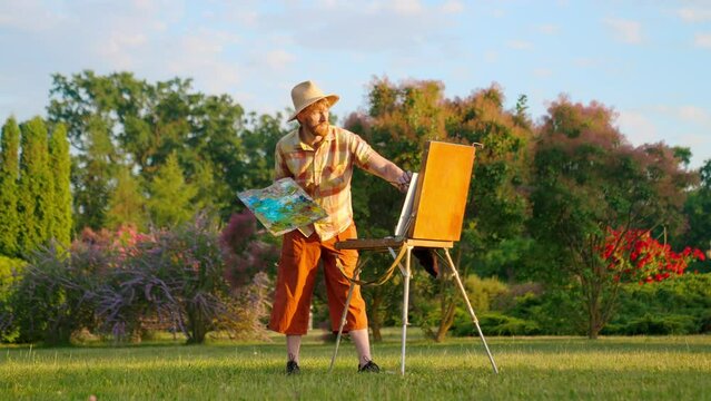 The artist paints a picture in the park. Green landscape, grass trees and flowers. The easel on which the painting stands. Blue, green colors in the palette.