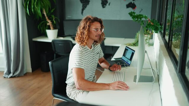 Smiling young curly long haired man in eyeglasses working on computer in office  